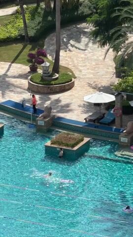 A resort swimming pool is shown from an elevated perspective. The pool is a vibrant turquoise color with visible lane markers on the bottom. Several people are swimming in the pool, some with swim caps. In the shallow end, a few individuals are relaxing on lounge chairs under umbrellas. One person stands near the pool's edge, possibly a lifeguard or staff member. In the background, there are palm trees, lush greenery, and architectural elements of the resort. The lighting suggests it is daytime.