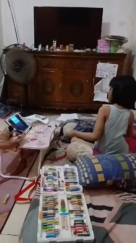 A young girl with black hair is lying on a bed in a room, facing a tablet that is propped up on a folding table. She is wearing a light blue tank top. In front of her on the bed is an open box of crayons, filled with rows of colorful crayons. Behind her, a large fan is visible, and further back is a wooden dresser with a television on top. The room appears to be a bedroom, with pillows and bedding spread across the bed. The girl is interacting with the tablet, her hands near the screen.