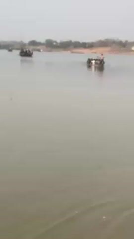 The video shows a wide, calm river with several small boats carrying people. The boats appear to be moving towards the opposite shore, which is visible in the background and is lined with trees. The water is a murky color, and the sky is overcast. There is also some footage of a wooden structure along the riverbank. Overall, the scene depicts a serene river crossing.