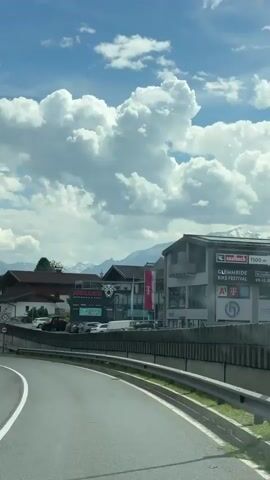 This video captures a view from a car driving through Mitterhofen, Austria. The scene is dominated by a bright blue sky filled with large, fluffy white clouds, suggesting pleasant weather, likely daytime. Towering mountains form a backdrop, their peaks possibly snow-capped, indicating a mountainous region.

Along the roadside, several buildings are visible. One building features signage for "HOLLIE'S SNOWMOBILE & QUADS," suggesting an adventure or recreational activity business. Another building has prominent branding for "T-Mobile" and appears to be a retail or service establishment. A sign for "GLEMMTAL BIKE FESTIVAL" is also visible, hinting at a local event or attraction.

Traffic signs are present, including a speed limit sign indicating 80 and a sign prohibiting trucks. The road itself is paved, with a concrete barrier separating the driving lane from the adjacent area. The overall impression is of a scenic drive through a small Austrian town or village, likely during the summer season given the presence of the bike festival advertisement and the lush greenery. The video conveys a sense of travel and exploration.