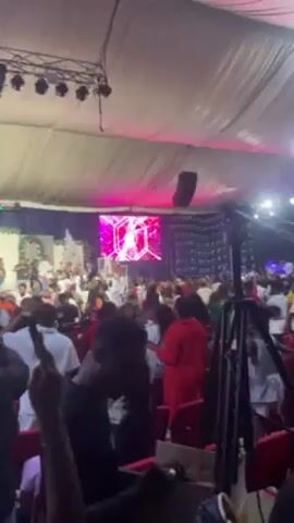 Here is a detailed description of the video.

The video captures a lively indoor event, possibly a concert or show, with a large crowd gathered in a large tent-like structure. A stage is visible at the front, decorated with a screen displaying a red hexagonal logo. Performers are seen on the stage, though their specific actions are obscured by the crowd.

Attendees are densely packed, some holding up phones to record the event, others gesturing or dancing. The environment appears to be well-lit, with stage lights casting various colors across the scene, including pink. The tent structure has a slightly off-white, almost beige, interior fabric. Some attendees are seated on red chairs. Security personnel are visible near the foreground.