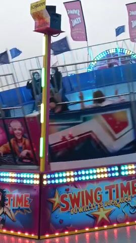 The video shows a carnival ride called "Swing Time." The ride is a large, spinning amusement with multiple cars that swing outwards. The ride is decorated with bright lights and colorful images. People are seen riding the attraction, with some appearing to be enjoying the experience while others are being tossed around. The background appears to be an outdoor setting, possibly a fairground or amusement park. There are flags with the ride's name displayed. The video is shaky, indicating it was likely filmed by someone on the ride or standing very close to it. The time stamp at the beginning of the video is 00:00 and it continues until 00:04.