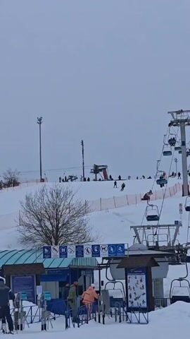The video shows a ski resort with a chairlift, a ski slope, and people skiing and snowboarding. There is a ticket booth and a few people standing around. The sky is overcast and the ground is covered in snow.  Someone has left their snowboard on the ground near the ticket booth.