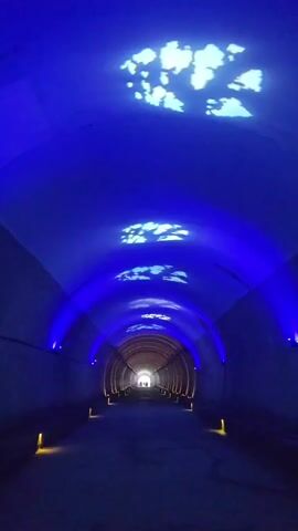 The video shows a tunnel with blue lighting and cloud projections on the ceiling. There are lights on the sides of the tunnel and at the entrance. There are people walking in the distance at the end of the tunnel. The tunnel appears to be made of concrete and has a curved ceiling.