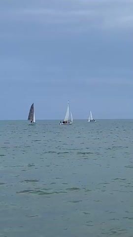 The video shows three sailboats on a calm sea under a cloudy sky. The sailboats are moving from right to left across the frame, with the closest boat on the left. Each sailboat has at least one person on board. The water is a deep blue-green, and the sky is a pale gray-blue with streaks of clouds. The video is a still shot of the scene.