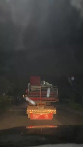 A car is driving at night on a rural road, following a flatbed truck carrying machinery. The truck has flashing yellow hazard lights. There are a few buildings and lights visible in the dark background. Another truck with headlights on approaches from the opposite direction. The windshield of the car is dirty.  Music is playing.