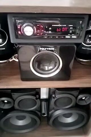 The video displays a close-up view of a sound system. The sound system consists of a car stereo head unit with a digital display showing "bt ON", a square black subwoofer with the brand "POLYTRON" printed on it, and two pairs of smaller speakers, all arranged on wooden shelves. The lighting suggests an indoor setting. The sound system is not in operation, as indicated by the static image and lack of audio.