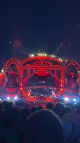 The video shows a crowd of people at an outdoor music festival at night. The stage is elaborately decorated with red and gold accents, and it is lit up with red lights and spotlights. The crowd is densely packed and appears to be enjoying the music, with many people holding up their hands. The sky is dark, with a few stars visible. There is also a haze of smoke or fog in the air, possibly from pyrotechnics or fog machines. The overall atmosphere is energetic and festive.