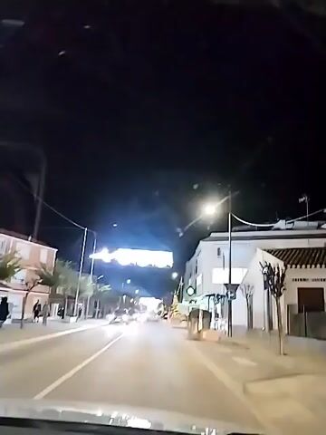 A driver films the road ahead at night through the windshield of a car. The car is traveling on a well-lit street. Several vehicles pass by in the opposite direction. On either side of the road are buildings with illuminated signs. A Christmas tree stands outside one of the buildings. Pedestrians walk on the sidewalks. Overhead, decorative lights are strung across the street.