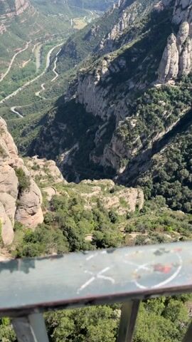The video presents a panoramic view of a mountainous landscape, likely captured on a sunny day. The foreground features rocky cliffs interspersed with lush green vegetation, creating a rugged terrain. Further down, a road snakes through a valley with a river. The perspective shifts upward to reveal towering rock formations characteristic of mountain ranges, dotted with trees and shrubbery clinging to the steep slopes. The scene is bathed in natural light under a clear blue sky, conveying a sense of height and the immensity of the environment.