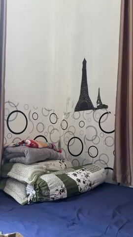 The video displays a corner of a bedroom. On the floor, there is a dark blue bedsheet covering a mattress. A stack of pillows and folded blankets rests on the bedsheet, leaning against the wall. The wall itself is painted white and decorated with a wallpaper featuring a pattern of overlapping black and white circles. Towards the top of the wall, two black silhouette decals of the Eiffel Tower are visible. On the right and left sides of the frame, brown curtains are partially drawn. The lighting appears to be artificial, creating soft shadows. There are no people or animals present in the video.