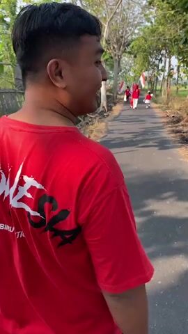 The video shows a person from behind, wearing a red t-shirt with white and black text and an emblem on the back. The text appears to read "INDONESIA" with an eagle emblem. The person is walking on a paved road, carrying a cardboard box in their left hand. The road is lined with trees and vegetation, and there is a bamboo fence on the left side of the road. In the distance, several people in red and white are walking away from the camera, some carrying Indonesian flags. The scene appears to be outdoors during daylight, with sunlight filtering through the trees. The camera follows the person from behind.