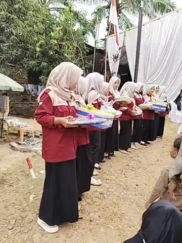 The video shows a group of young women, dressed in maroon shirts and black skirts with headscarves, standing in a line. Each woman is holding a gift-wrapped box or tray containing what appears to be food items, possibly for a celebration or event. They are all facing towards the right side of the frame. In front of them, a young boy, wearing a brown shirt and black pants, is reaching out towards the line of women. The background features a white tent with draped fabric, palm trees, and some debris on the ground, suggesting an outdoor event. The overall scene appears to be an offering or distribution of gifts.