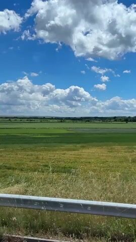 The camera captures a scenic view of a landscape from behind the passenger seat window of a vehicle moving along a road. The sky is predominantly blue with scattered, fluffy white clouds. Below the sky, a vast expanse of green and golden fields stretches out to the horizon. A grey metal guardrail runs along the edge of the road, separating the vehicle from the fields. Patches of roadside grass and a few scattered trees dot the landscape. In the distance, a range of hills can be seen. The overall impression is one of a rural or agricultural area on a bright, sunny day.