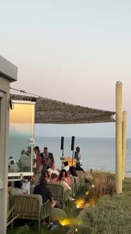 A vertical video captures an outdoor gathering near the sea at dusk. In the foreground, to the left, is a portion of a building with a mirrored surface reflecting people. To the right, a grassy slope leads down towards the ocean. Several groups of people are seated on outdoor furniture, facing a stage area under a shaded canopy. On the stage, two speakers flank a podium where a person appears to be speaking or performing. The sea is visible in the background, with a sandy beach and some structures further down the coast. The sky is a gradient of soft colors, indicating sunset. The overall atmosphere suggests a relaxed, social event.