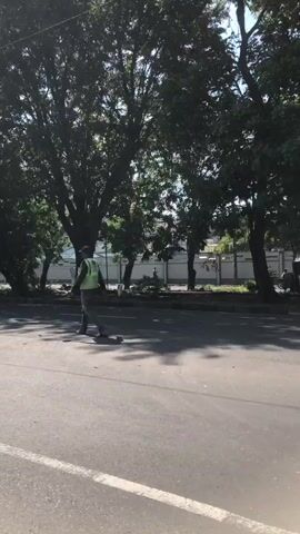 The video shows a man in a reflective vest and dark pants walking across a paved road. He is facing away from the camera and appears to be moving from left to right. Behind him, on the side of the road, there are trees and some buildings or fences. Two other people are visible on the sidewalk or grass area behind the walking man, but their actions are unclear. The lighting suggests it is daytime.