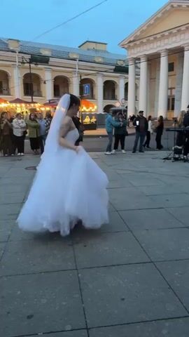 A person in a white wedding dress with a veil spins around in a city square. There are other people in the background, some standing and watching, and a band playing music. The buildings in the background appear to be old and ornate. The sky is a dusky blue, suggesting it is either early morning or late evening.
