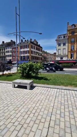 The video opens on a cobblestone path with a concrete bench on the left, a grassy area, and multi-story buildings in the background under a clear blue sky. The camera pans right, revealing large white letters spelling "MACON" on a green median. A street is visible behind the letters with a black van and a scooter driving. The camera continues panning right, showing a paved walkway where a person on a bicycle and another walking individual are present. The pan concludes with a view of a wide body of water, partially obscured by a green hedge, and a multi-arched bridge in the distance.