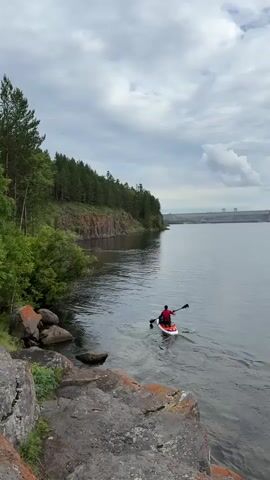 A person in a red shirt and black shorts is paddleboarding on a calm lake. The paddleboarder is facing away from the camera and is paddling towards the right side of the frame. The lake is surrounded by trees and rocky cliffs. In the background, there is a large dam. The sky is cloudy.