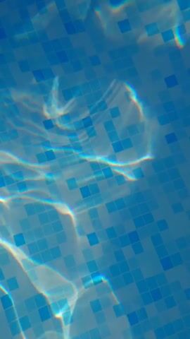 The video shows an overhead view of the tiled bottom of a swimming pool. Light refracts and dances on the surface of the water, creating shifting patterns of brightness and distortion over the blue and white tiles. There are no people or objects visible in the pool, and no actions are taking place other than the natural movement of the water.