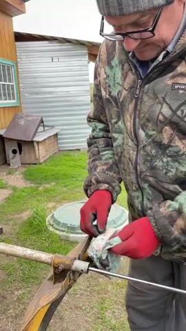 A person wearing red gloves is using a cloth to clean a metal rod that is connected to a yellow, blade-like object attached to a rusty, metal pole. The person is wearing a camouflage jacket and grey pants, and black shoes. The person is standing on grass, and a metal building, dog house, and sewer access point are visible in the background. The person moves the cloth around the rod and the connected part to clean it.