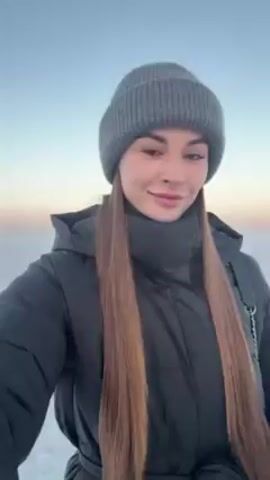 A young woman is standing in a snowy landscape. She is wearing a dark grey puffer jacket with a tie at the waist, black leggings, grey knitted socks, and a grey beanie. She has long, straight brown hair that falls over her chest. She is also wearing a small black purse slung over her shoulder. The background is a flat expanse of snow under a light blue sky that transitions to pink on the horizon. The woman looks at the camera and smiles. She then turns to her right and looks out over the snow. The scene is brightly lit by natural light.