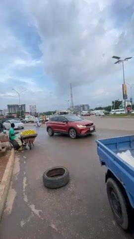The video shows a street scene with moderate traffic. In the foreground, on the left, a vendor is pushing a cart laden with oranges. A few people are standing near the cart. In the middle of the frame, a white sedan is stopped in traffic. Behind it, a dark sedan is also stopped. To the right, a red SUV is moving. Further down the street, a large tanker truck with "NIPCO" written on its side is visible. Various other vehicles, including cars and trucks, are present on the road. Several people are seen walking, including one crossing the street. Buildings and a tall communication tower are in the background. The sky is cloudy, suggesting overcast weather. The road surface appears wet in some areas.