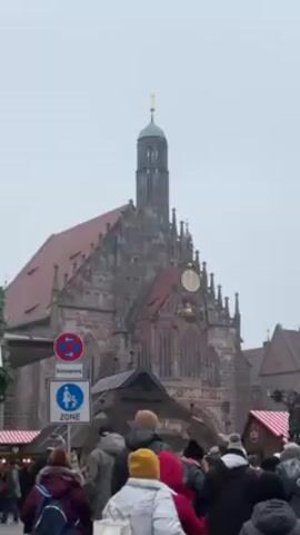 The video shows the Frauenkirche church and the Schöner Brunnen fountain in Nuremberg, Germany, surrounded by a crowd of people. The Frauenkirche is a large Gothic church with a red tile roof and a tall, slender tower. The Schöner Brunnen is a tall, ornate fountain with many figures and sculptures. The sky is overcast, and the scene appears to be a bustling market or gathering place.