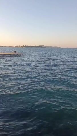 This video captures a serene coastal scene in Urla, Turkey, likely during the late afternoon or early evening as suggested by the soft, warm light. The primary subject is the vast expanse of the sea, with gentle ripples creating a textured surface that reflects the clear, pale blue sky. In the distance, a thin strip of land, possibly an island or the mainland coast, stretches across the horizon, with subtle outlines of buildings or structures visible. The overall atmosphere is peaceful and tranquil, evoking a sense of calm and natural beauty. There are no people or specific objects prominently featured, allowing the viewer to focus on the expansive seascape and the subtle interplay of light and water. The location cues, such as the distant landmass and the characteristic Turkish coastal light, strongly suggest Urla, Turkey, as the setting. The weather appears to be pleasant, with calm waters and a clear sky. There is no visible text within the media file.