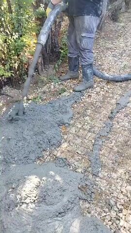 A man is pouring wet concrete from a hose onto a grid of metal mesh on a gravelly surface. He is wearing work clothes, including grey pants, a dark t-shirt, and black boots. Another man is in the background, also holding a hose and directing concrete. The environment appears to be outdoors with trees and vegetation. The concrete is being laid to form a path or foundation.