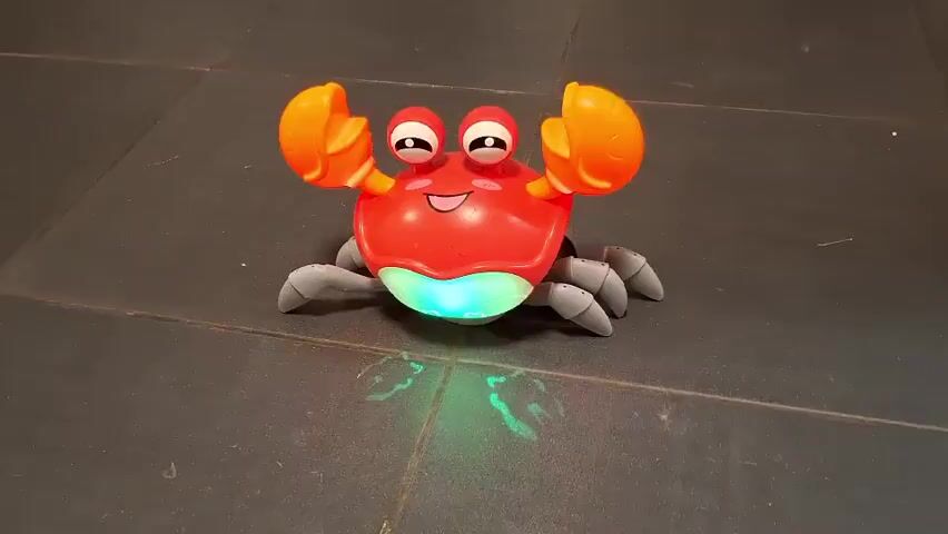 A red toy crab with orange claws and gray legs sits on a dark tiled floor. The crab has large eyes with black pupils and white highlights, and a smiling mouth. Its underside glows with a soft green light. As the video progresses, the crab moves slightly, its legs shifting and the green light underneath it pulses. The toy casts a faint green shadow on the floor, mimicking the shape of crab legs. The camera remains stationary, focusing on the toy crab as it performs its programmed movements and lights up.