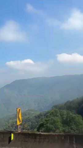 The video captures a scenic view of mountains and a blue sky with clouds. In the foreground, there is a concrete wall or barrier. Lush green trees and vegetation cover the hillsides. In the distance, a viaduct or bridge can be seen spanning across the valley. The camera pans slightly, revealing more of the landscape and some road signs. The lighting suggests it is daytime with bright sunlight. There are no people visible in the video.