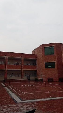 The video shows an outdoor badminton court during a rainfall. The court, marked with white lines on a wet red surface, is situated between a red brick building and a volleyball net. Several people can be seen standing on a balcony of the building, observing the scene. The benches surrounding the court are unoccupied. The sky overhead is overcast.