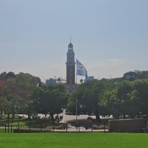 The video shows a cityscape with a large clock tower and the Argentinian flag in the foreground. The scene is framed by a green lawn and a black fence. Trees dot the landscape, and there are a few people walking in the distance. In the background, buildings and construction cranes can be seen. The sky is clear and bright.