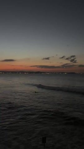 The video shows a dark ocean scene at dusk with a colorful orange and gray sunset visible on the horizon. There are a few sparse clouds in the sky. The water is dark, with visible waves. Two surfers are visible in the water. In the distance, a city skyline is lit up with lights. The bottom of the frame is cut off with a black shadow.