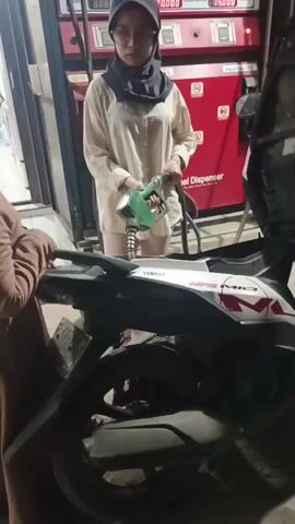 A young woman wearing a beige hijab and a cream-colored long-sleeved shirt is holding a green gasoline nozzle and appears to be refueling a white Yamaha scooter. She is standing at a red gas pump with digital displays. To her left, another young girl, wearing a brown outfit and a matching brown hijab, is looking at the scooter. The scene takes place at a gas station at night, with the pump and scooter illuminated by artificial light.