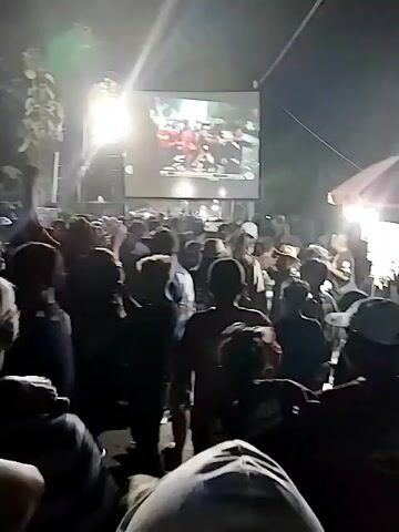 A large crowd of people are gathered outdoors at night, watching a large screen that is displaying a video. The screen is illuminated by bright lights, and the crowd is silhouetted against the light. Some people in the crowd are raising their hands in the air, and others appear to be dancing. The atmosphere seems festive and energetic. The video on the screen is difficult to discern due to the image quality, but it appears to show multiple people, possibly on a stage. The environment appears to be a natural setting with trees visible in the background. There are also some tents or structures present.