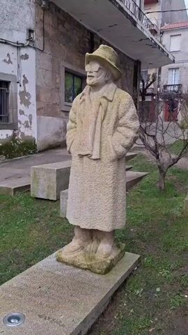The video shows a statue of a man in a hat and overcoat standing on a stone pedestal in a grassy park. The statue is made of a light-colored stone and is weathered, with some green algae or moss growing on it. The man is wearing a hat, glasses, and a long overcoat with his hands in his pockets. A scarf is also draped around his neck. The pedestal has writing etched into it. The park is surrounded by buildings with stone facades and small trees. The sky is overcast, suggesting a cloudy day.