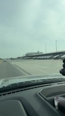 The video shows a view from inside a vehicle, looking out the windshield. The vehicle is on a paved surface, likely a road or runway. To the right, a long row of identical light-colored vehicles is parked in parallel formation. These vehicles appear to be cars or SUVs. In the distance, a low, long building with a white facade is visible, with some taller structures on top. Several lampposts are spaced along the parking area. To the left, other vehicles are seen on a roadway, moving away from the camera. The sky is overcast with a light gray hue. The dashboard of the vehicle is visible in the foreground, with a dark, textured surface. A black object, possibly a dashcam or mount, is attached to the windshield in the upper right corner.