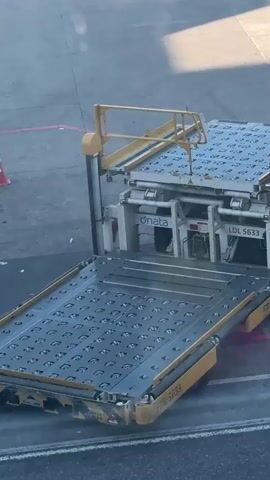 A high-angle view shows an airport tarmac with a person standing on top of a yellow scissor lift next to an airport cargo loader. The cargo loader has two platforms, one lower and one higher, both covered with a pattern of circular metal studs. The loader has "dnata" and "LDL 5633" written on its side. The tarmac is gray with yellow and red painted lines. The sun casts a shadow from the loader and the person.