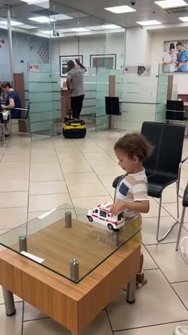 A toddler boy with curly brown hair is playing with a toy ambulance on a glass coffee table in what appears to be a waiting area. He is wearing a striped shirt and yellow shorts. In the background, a man is standing on a yellow cart and working on something above him. A woman is seated at a desk in the left background, and there are other chairs and frosted glass partitions. The floor is tiled.