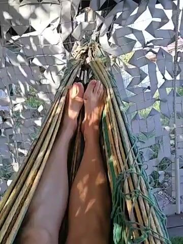 Two bare legs are visible in a hammock, with the person's feet at the top of the frame. The legs are tanned, with one leg appearing slightly darker than the other. The hammock is made of woven material and appears to be suspended outdoors. The background consists of greenery and a camouflage-like netting. The video shows slight movement, suggesting the hammock is gently swaying.