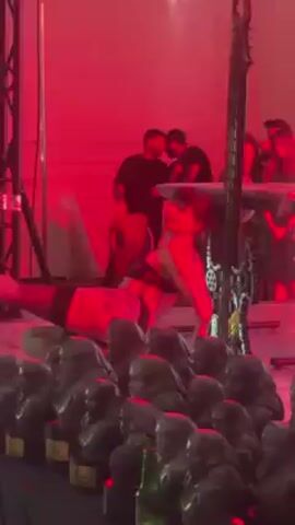 The video shows a row of trophies in the foreground with an indistinct group of people and equipment in the background. The trophies appear to be bust sculptures of people's heads. In the background, several people are standing and appear to be operating equipment that includes a keyboard and an electrical instrument. A performer wearing shorts and black stockings is doing a handstand in front of the trophies. The scene is lit in red light.