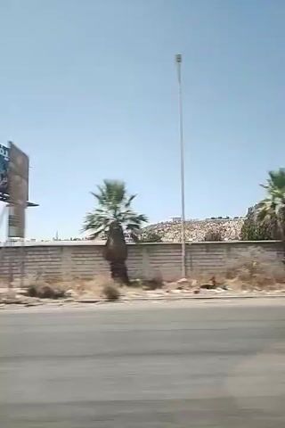 The video shows a view from a moving vehicle on a sunny day. On the left, a large billboard advertising what appears to be a travel destination is visible. Beyond the billboard, there are palm trees and a low brick wall. In the distance, a hillside dotted with structures or graves is discernible. To the right of the billboard, a street light stands tall. Further to the right, the scene continues with more palm trees, a brick wall, and in the far distance, a densely populated hillside. The road in the foreground shows motion blur, indicating the perspective of a moving camera. There are no people or specific actions clearly depicted. The environment appears arid with dry vegetation alongside the road.