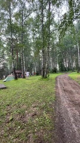 A person in a white shirt and shorts runs across a grassy area with trees. There are several tents and small structures in the background, and a dirt path curves through the trees to the right. The overall scene appears to be a campsite in a wooded area.