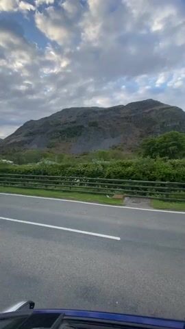 The video shows a drive along a road through a countryside with hills, green vegetation, and a cloudy sky. The car stops. A young man dressed in a tracksuit gets out of the car and looks at his phone. A young woman then emerges from the sunroof. She leans out and looks at the camera.