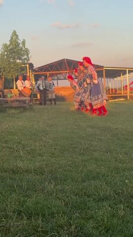 This is a video of a cultural performance taking place outdoors in Voynegovtsi, Bulgaria. The scene depicts a group of women in traditional Bulgarian folk costumes, characterized by blue dresses with intricate white embroidery and red headscarves, performing a synchronized dance. They are accompanied by musicians playing traditional instruments, including an accordion and a drum.

The setting appears to be a rural area, with a grassy field in the foreground and a rustic wooden structure with a thatched roof in the background, possibly a stage or seating area. A few other people are visible, some seated and others standing, observing the performance. The sky is a pale blue with scattered clouds, and the warm, soft lighting suggests it is either early morning or late afternoon, likely during sunset, given the golden hue of the light. The overall atmosphere seems festive and communal, with the vibrant costumes and lively music contributing to a cheerful mood. There is no visible text in the media.