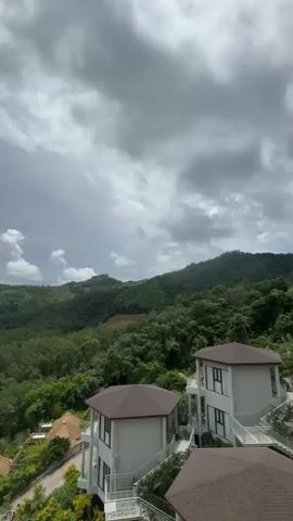 A vertical video shows a drone flying over a green, hilly landscape under a cloudy sky. In the foreground, several modern houses with brown roofs and white walls are visible, some with balconies and railings. A road winds through the houses. Beyond the houses, dense green trees cover the rolling hills and mountains. The sky is filled with dramatic, gray and white clouds, with some patches of blue sky and sunlight breaking through. In the distance, islands can be seen in a body of water. A yellow caution sign with text in an unknown language is placed on the ground near a railing in the foreground. The drone appears to be moving forward and upward, gradually revealing more of the landscape.