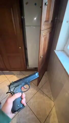 The video shows a person holding a black pistol in their right hand. The person first holds the gun in front of an open cabinet with a mirror inside. Then, the person moves the gun to an open window where there are white buildings and grass visible outside, along with a slightly cloudy sky. The person slowly rotates the gun in front of the window.  The music plays dance music in the background.