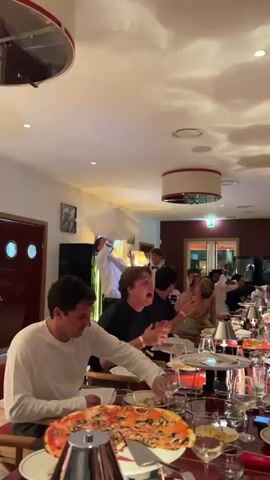 A group of people are seated at a long table in a restaurant. A man in a white shirt is standing at the head of the table and tossing pizza dough into the air. People at the table are looking up at the dough and some are smiling and holding up their phones to record. There are pizzas on the table. The room is dimly lit and decorated with modern fixtures and artwork.